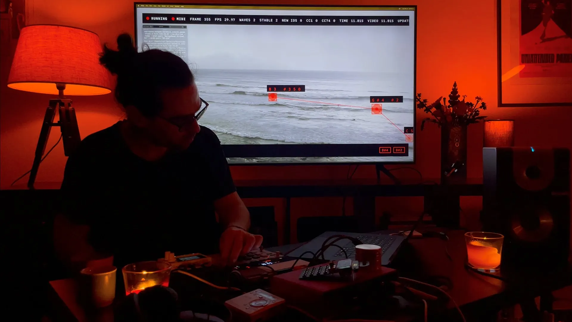 At the synthesizers in a red-lit room, the beach cam's tracked waves on the screen behind